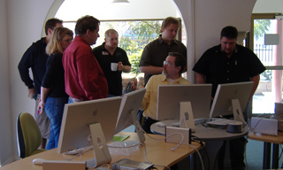 A group of people standing and looking at computer monitors in an office setting.