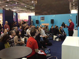 An audience sits in rows listening to a man in a red shirt speaking at a convention booth.
