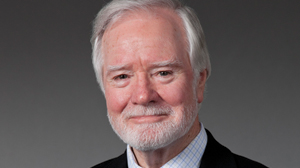 An older man with white hair and a white beard smiling against a gray background.