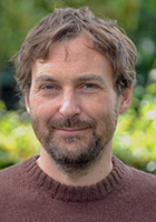 A man with brown hair and a beard wearing a brown sweater, smiling against a blurred green background.