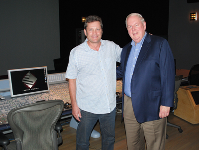 Two men smiling and standing together in a professional recording studio next to a mixing console.