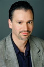 A man with dark hair and a goatee wearing a grey blazer and dark shirt against a grey background.
