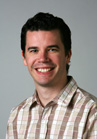A man with dark hair smiling while wearing a tan and white plaid button-down shirt.