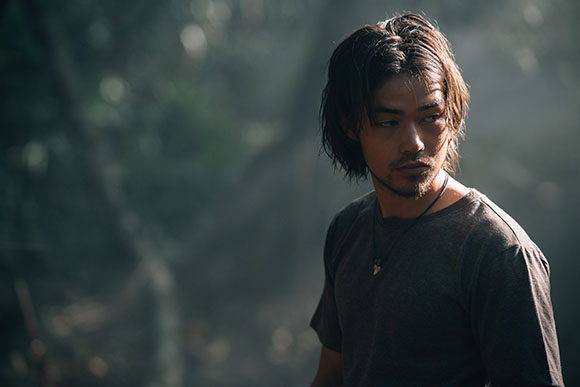 A man with medium-length dark hair and a beard looking to his right in a dimly lit forest.