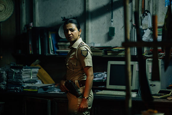 A woman in a tan police uniform stands in a dark, cluttered office holding a handgun.