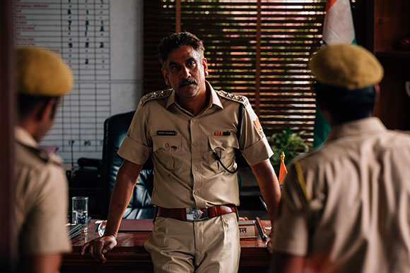 A police officer in tan uniform stands behind a desk, facing two other officers in an office.