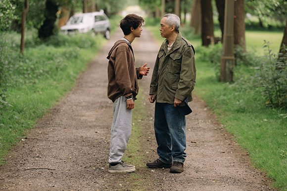 A young man in a brown hoodie and grey sweatpants talks to an older man in an olive jacket on a park path.