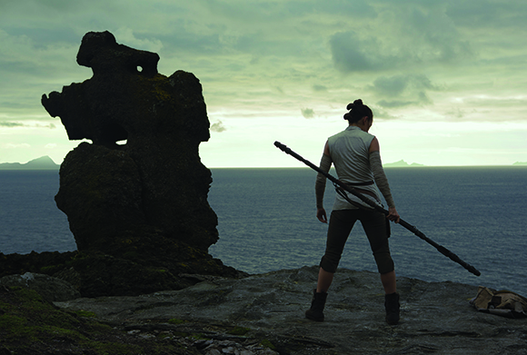 A person in light clothing holds a long staff, standing on a rocky cliff facing the ocean.