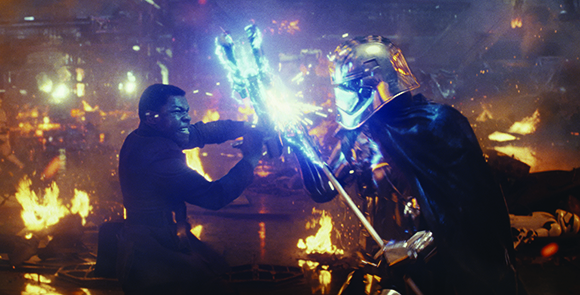 A man in dark clothing and a figure in a silver helmet clash lightsabers in a fiery, dark environment.