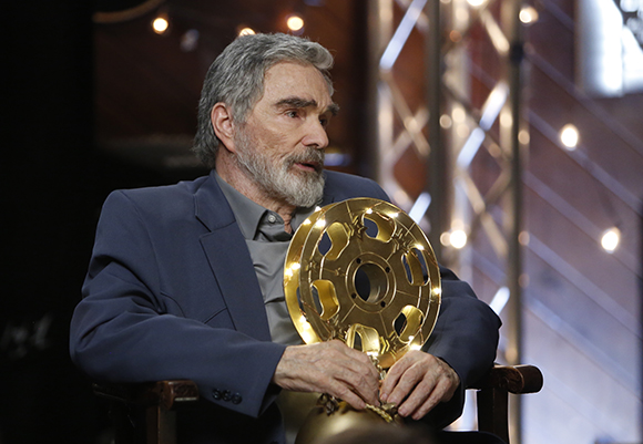 A man with gray hair and a beard sits in a chair holding a large gold circular award.