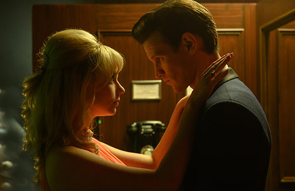 A blonde woman in a pink dress looks up at a man in a dark suit in a dimly lit room.