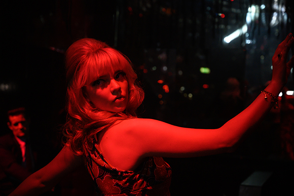 A blonde woman in a patterned dress poses under intense red lighting in a dark setting.