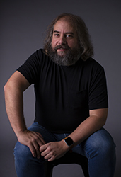 A man with long brown hair and a full grey-flecked beard wearing a black t-shirt and blue jeans.