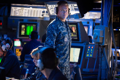 A man in blue camouflage military uniform wearing a headset stands in a dimly lit command center.