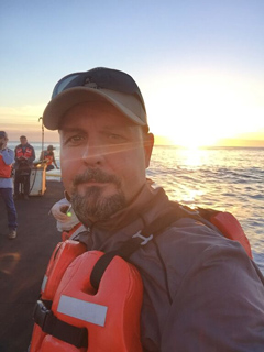 A man wearing a life vest and cap looks toward the camera on a boat during a bright sunset.