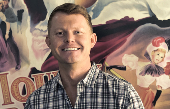 A smiling man wearing a blue and white plaid shirt stands before a colorful vintage-style mural.