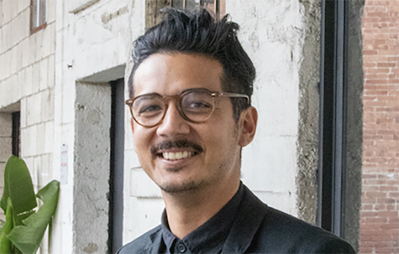 A man with dark hair, glasses, and a mustache smiling in front of a white stone building.