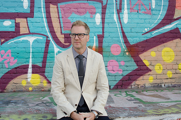 A man wearing glasses and a light blazer stands before a colorful graffiti-covered brick wall.