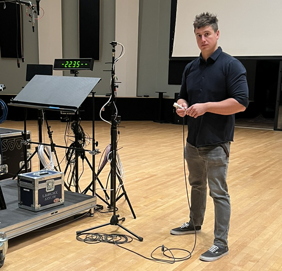 A man in a dark shirt and grey pants stands next to a music stand and audio equipment on a wooden floor.