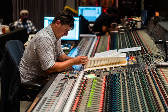 A man in a patterned shirt sits at a massive professional audio mixing desk, reviewing sheet music.