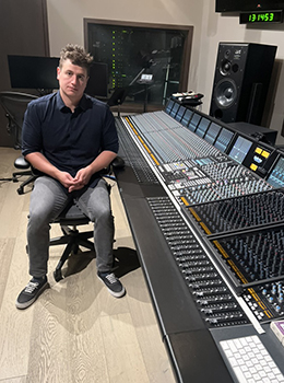 A man in a dark polo and grey pants sits in a chair beside a large professional audio mixing console.