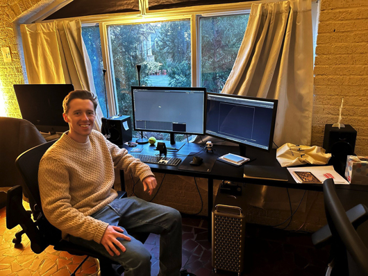 A man in a beige sweater sits at a desk with two computer monitors and a window in the background.