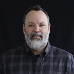 A man with short dark hair and a full grey beard wearing a dark plaid shirt against a black background.