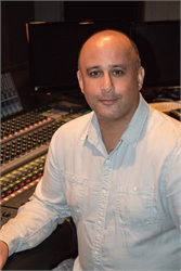 A man in a light blue button-down shirt sitting in a studio with a mixing console and monitors.
