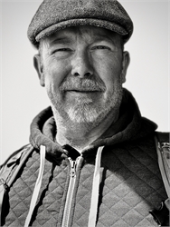 Black and white close-up portrait of a smiling man wearing a flat cap and a quilted zip-up jacket.