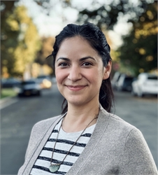 A smiling woman with dark hair wearing a striped shirt and a grey cardigan stands on a street.