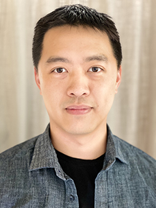 A man with short dark hair wearing a grey button-down shirt over a black shirt.