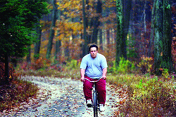 A man in a light blue shirt and red pants rides a bicycle down a leaf-strewn path in a forest.