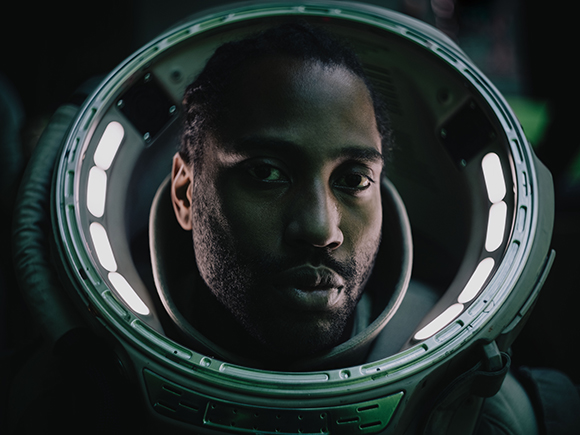 Close-up portrait of a man looking forward through the visor of a glowing space helmet.