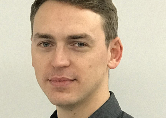 A close-up portrait of a man with short brown hair and blue eyes looking forward against a plain background.