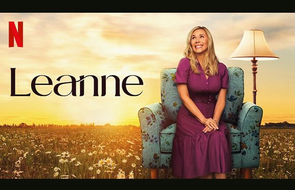 A smiling woman in a purple dress sits in a floral armchair in a field of daisies at sunset.