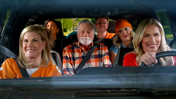 Six diverse people smiling while sitting inside a car, viewed from the dashboard perspective.