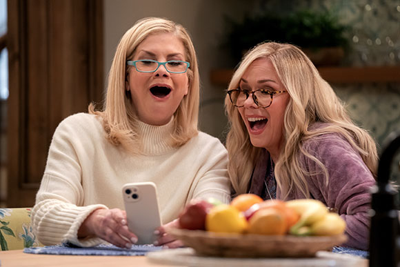 Two blonde women wearing glasses look at a smartphone with wide mouths and excited expressions.