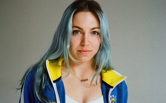 A woman with long, light blue hair and a blue and yellow zip-up jacket looks toward the camera.