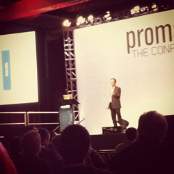 A man in a suit stands on a stage presenting to an audience. A large screen behind him displays the word 'prom'.