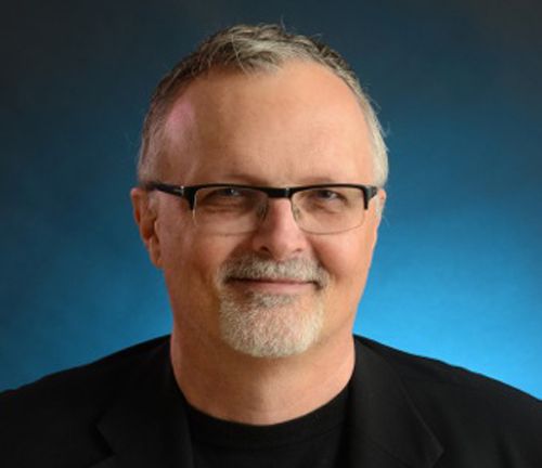 A man with short graying hair, glasses, and a gray goatee smiling against a blue background.