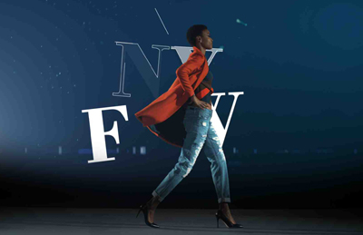A person in a red blazer and jeans walks past large white NYFW letters against a dark blue background.