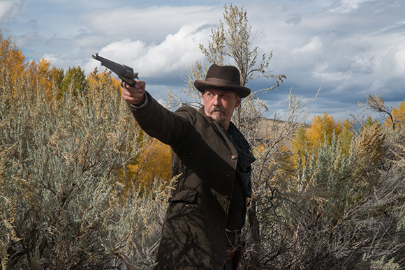 A man in a brown hat and coat points a handgun toward the left in a field of dry brush.