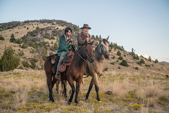 Two men in period clothing ride horses across a grassy, hilly terrain under a clear sky.