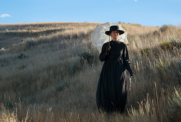 A woman in a long black dress and wide-brimmed hat holds a white lace parasol in a dry, grassy field.