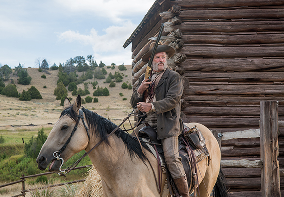 A bearded man in a cowboy hat and brown jacket sits on a tan horse in front of a rustic log cabin.