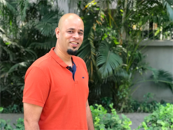 A bald man with a goatee wearing an orange polo shirt, smiling in front of lush green tropical foliage.