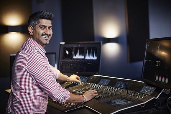 A smiling man in a red checkered shirt sits at a large professional audio mixing console with multiple monitors.