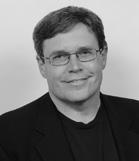 Black and white portrait of a smiling man with short hair and glasses wearing a dark blazer.