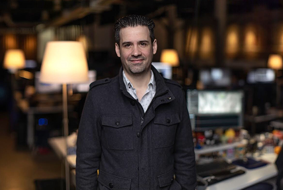 A man in a dark grey jacket smiling in a blurred office setting with lamps and computer monitors.
