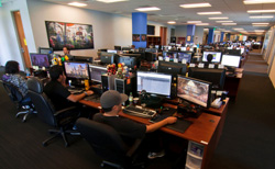 People sitting at long desks with multiple computer monitors in a large, open-plan office space.